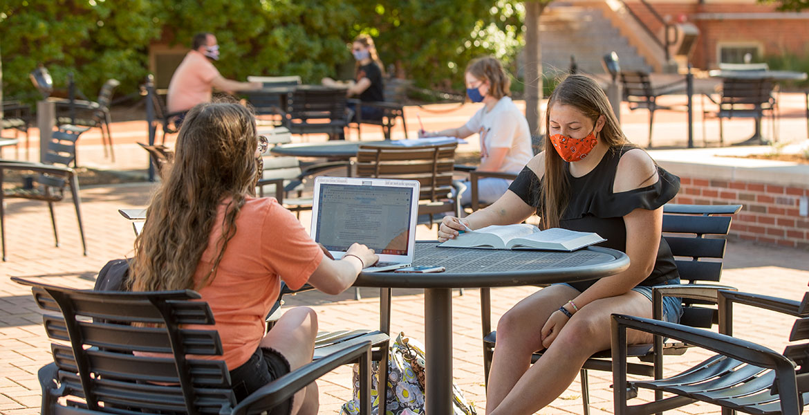 Two masked students sit an outdoor table and study.