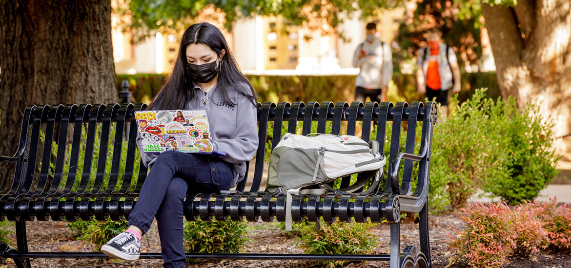 A masked student sits on a campus bench and looks at a laptop, which is decorated with colorful stickers covering the back.