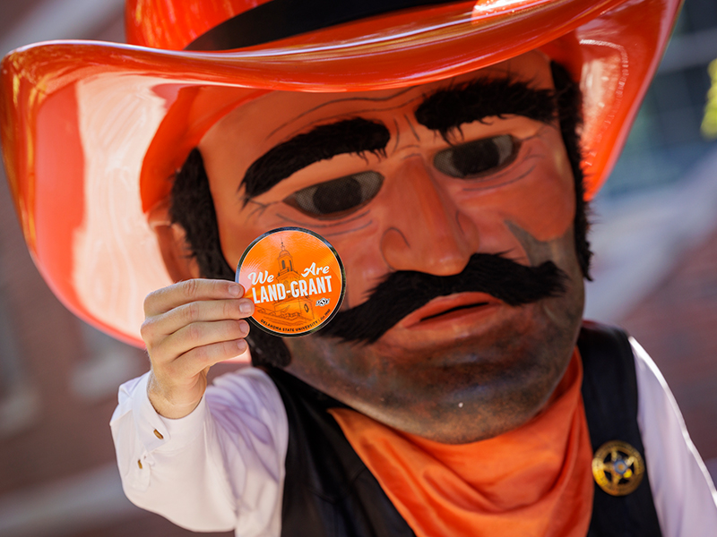 Photo of Pistol Pete holding up a sticker that says "We are Land Grant"